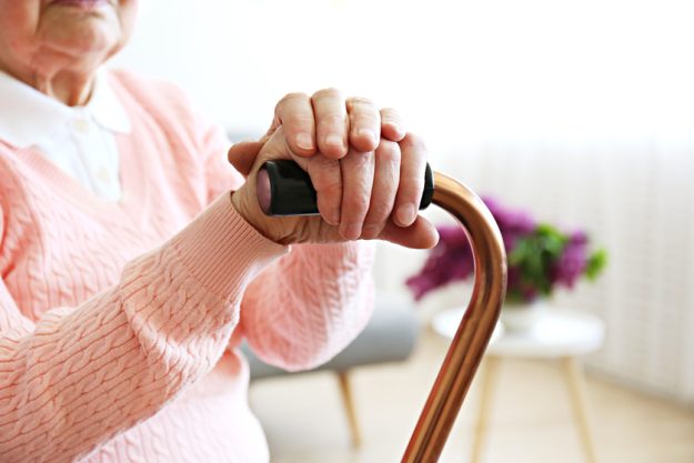 Elderly woman sitting in nursing home room holding walking quad cane with wrinked hand. Old age senior lady wearing beige cardigan, metal aid stick handle bar close up. Interior background, copy space A Guide To A Safe Living Environment For Your Aged Mom With Dementia