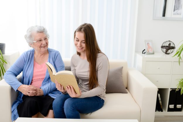 cheerful young woman reading a book for elderly senior woman at home