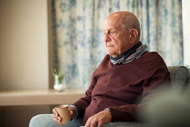 Depressed senior man sitting on armchair holding disposable cup of coffee and thinking. Frustrated retired man sitting on sofa and drinking a cup of hot tea. Sad mature man sitting alone at nursing home with sad expression. Depressed senior man sitting on armchair holding disposable cup of coffee and thinking. Frustrated retired man sitting on sofa and drinking a cup of hot tea. Sad mature man sitting alone at nursing home with sad expression.