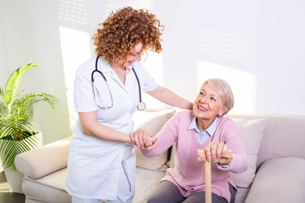Female caregiver helping senior woman get up from couch in living room. Smiling nurse assisting senior woman to get up. Caring nurse supporting patient while getting up from sofa